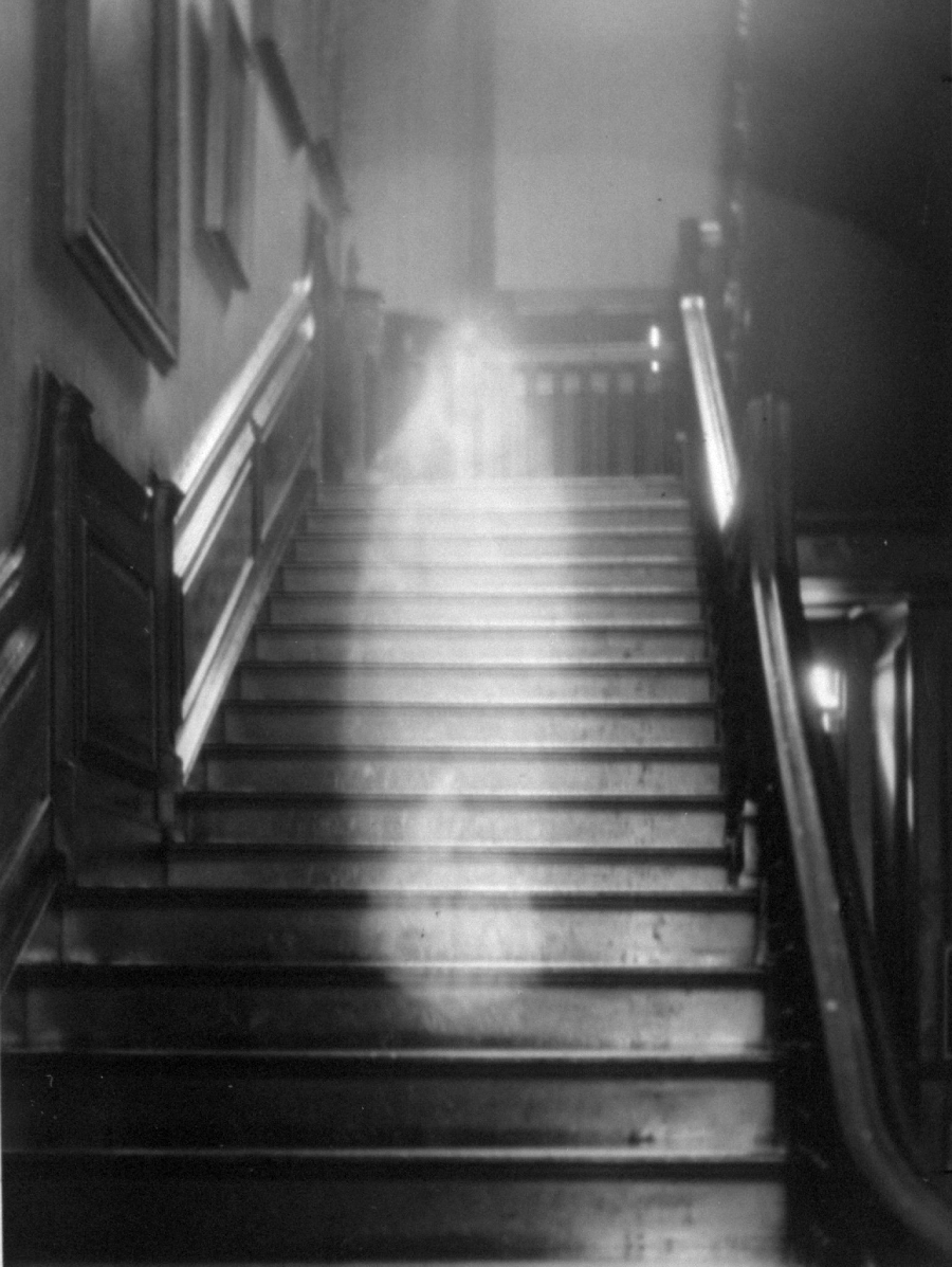 A black and white photo of a wooden staircase with a misty, semi-transparent white shape appearing in the middle of the steps, suggesting a ghostly or supernatural presence in an old, dimly lit building.