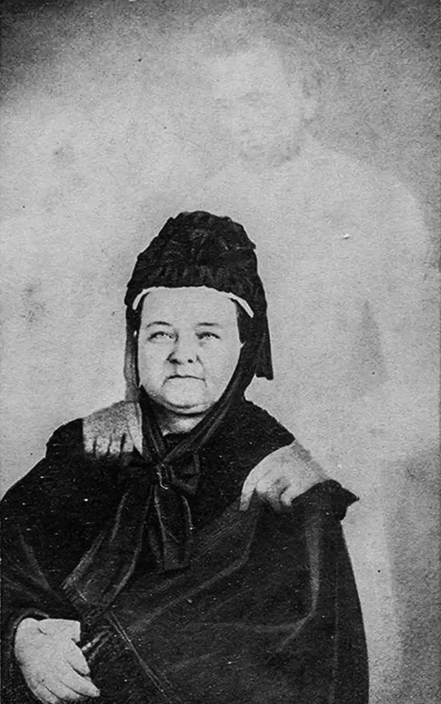 An older woman sits facing the camera, wearing dark clothing and a bonnet, with the faint, ghostly image of a man standing behind her, appearing to rest his hands on her shoulders.