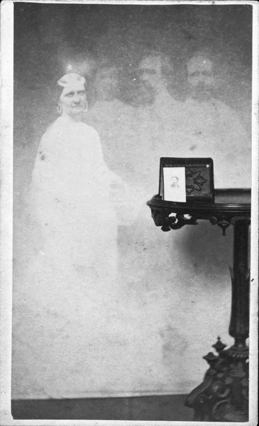 A black and white vintage photograph shows a faint, ghostly figure of a smiling woman in white beside a dark wooden table holding two framed photos. The image has a hazy, double-exposed effect.