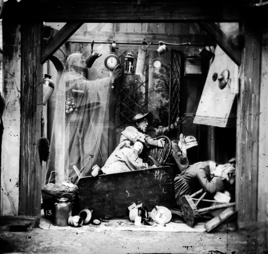 A black-and-white photo shows a chaotic scene: two frightened people cower amidst broken furniture and objects, while a ghostly, hooded figure looms in the background holding a scythe under a clock.