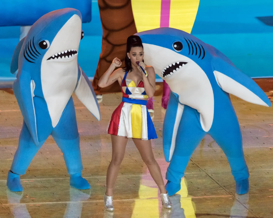 A singer in a colorful dress performs onstage, flanked by two dancers in blue shark costumes, against a bright, playful background.
