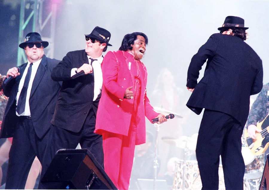 Three men in black suits, hats, and sunglasses perform on stage with a man in a bright red suit holding a microphone, singing energetically. Bright stage lights and musicians are visible in the background.