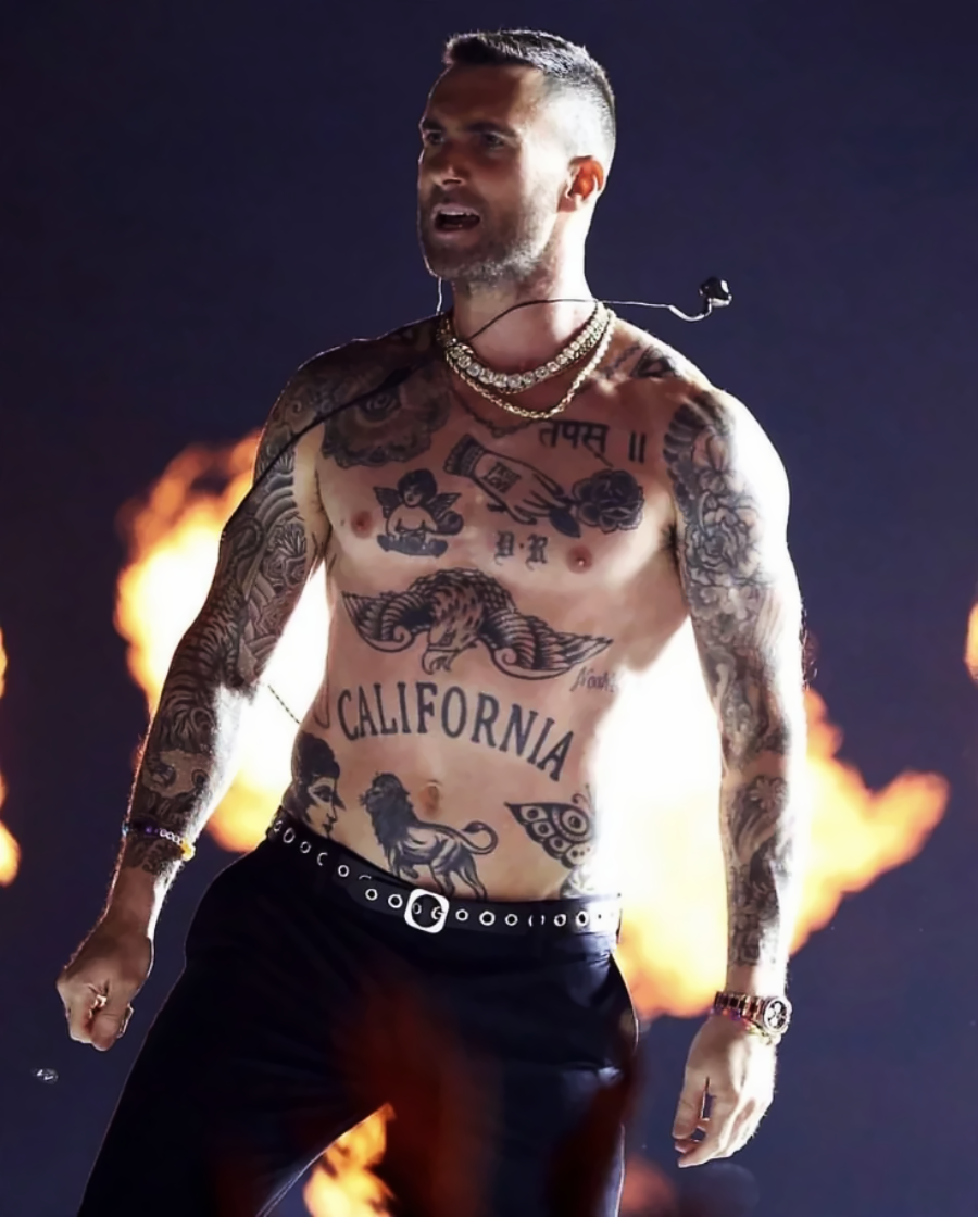 A shirtless man with tattoos performs on stage in front of large flames, wearing a chain necklace, belt, and black pants, with a microphone headset on.