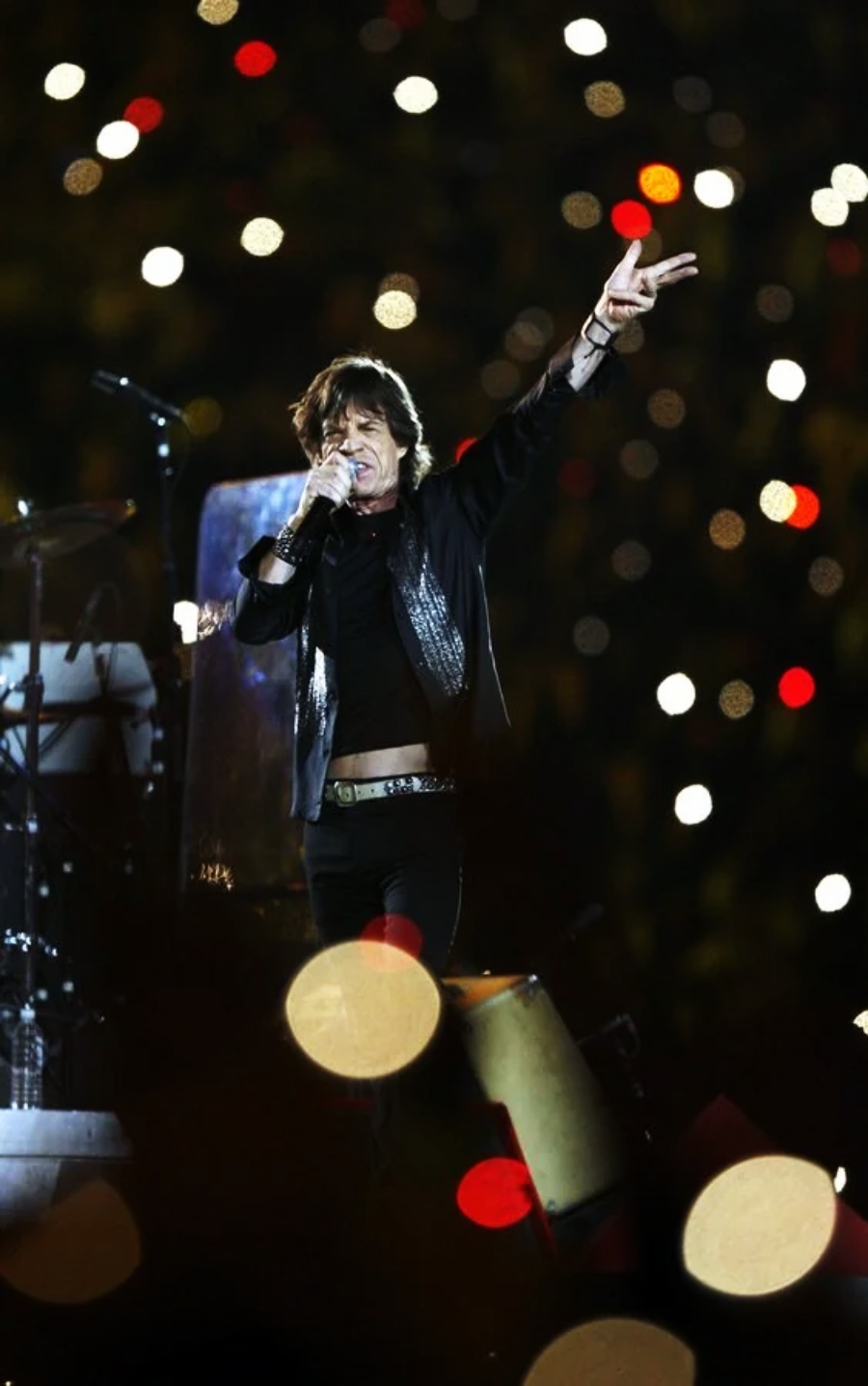 A rock singer performs energetically on stage, holding a microphone and raising one arm with fingers in a peace sign gesture. Blurred, colorful lights fill the background, creating a lively concert atmosphere.