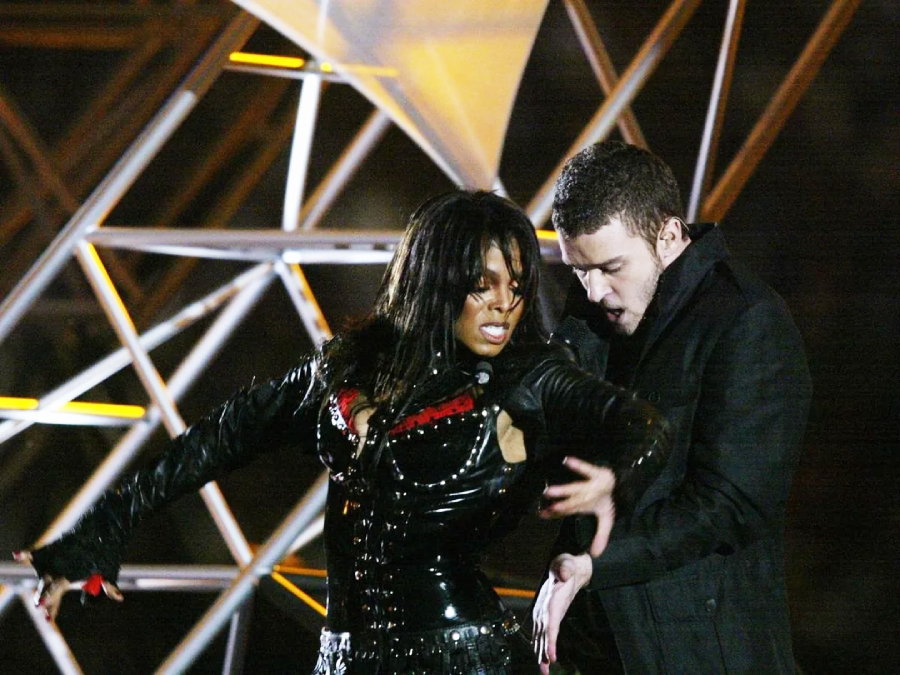 Two performers on stage during a high-energy show. One is wearing a shiny black outfit while the other stands close behind, reaching towards the first performer. A lit geometric structure is in the background.