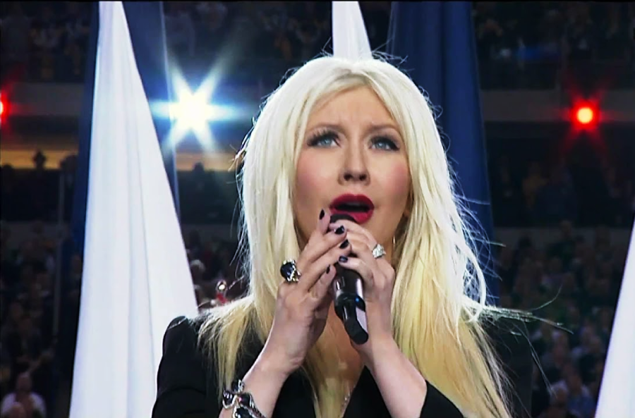 A woman with long blonde hair sings into a microphone in front of large flags, with bright lights shining in the background and an audience visible behind her.