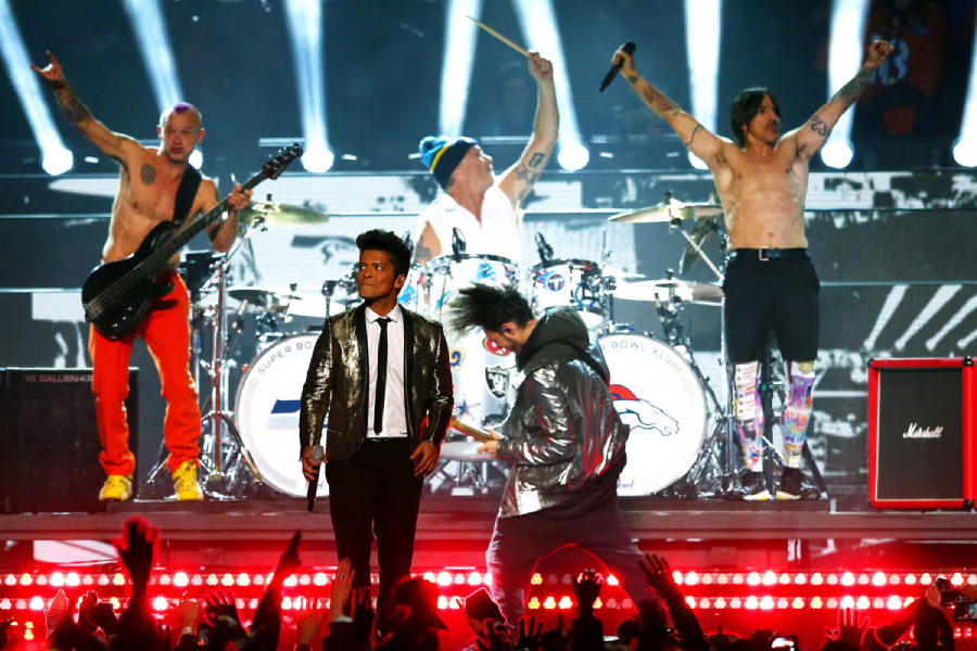 A band performs energetically on stage with two shirtless musicians, one playing bass and the other on drums, while two others in silver jackets sing and play guitar. Bright lights and a crowd with raised hands are visible.
