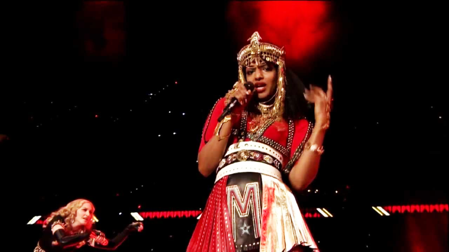 A performer in a red and gold costume, adorned with elaborate gold jewelry and a crown-like headpiece, sings on stage under red lighting. Another performer in black is visible in the background.