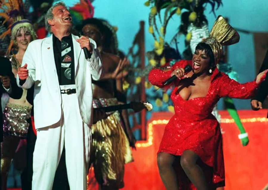 A man in a white suit and a woman in a sparkling red dress with a large gold headpiece sing and perform joyfully on stage, surrounded by dancers in colorful costumes.