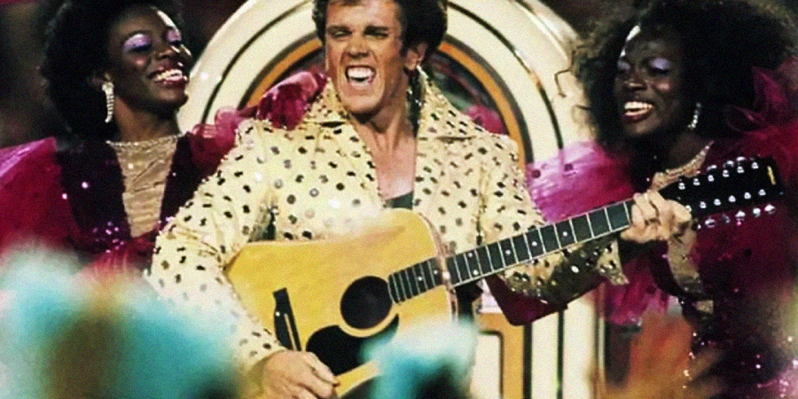 A man in a gold, sequined jacket enthusiastically plays an acoustic guitar and sings, flanked by two women in glamorous, sparkly outfits with bright pink ruffles, all performing on stage in front of a large jukebox.