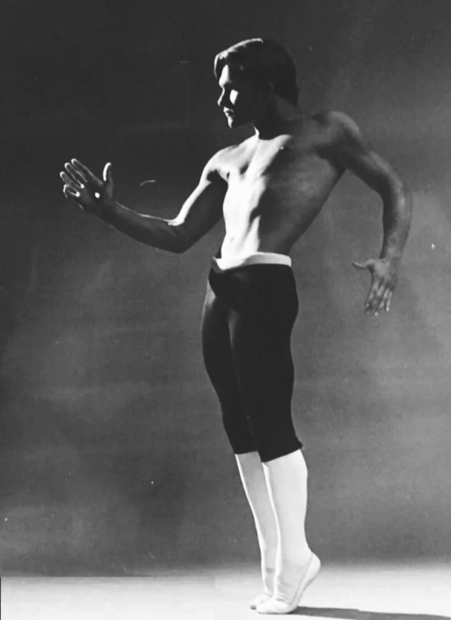 A shirtless dancer in dark tights and white socks poses gracefully, extending one arm forward and the other back, on a plain background. The lighting highlights his muscular form and expressive stance.