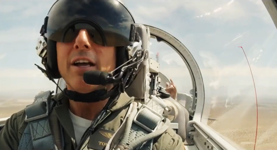 A pilot wearing a helmet and sunglasses sits in the cockpit of a jet, flying high above a desert landscape under a clear sky. The view shows cockpit controls and another person seated behind the pilot.