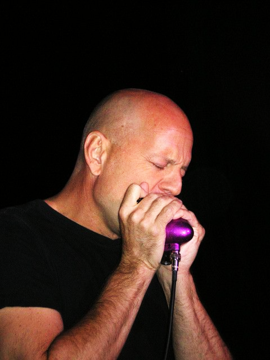 A bald man in a black t-shirt plays a purple harmonica into a microphone, eyes closed and focusing intently, against a dark background.