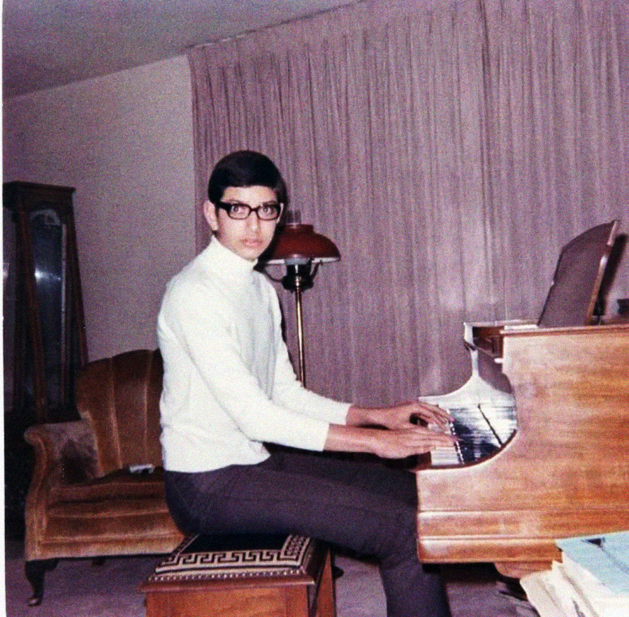 A young person with short dark hair, glasses, and a white turtleneck sits at a piano in a living room, looking toward the camera. The room has a brown sofa, a floor lamp, and closed curtains in the background.