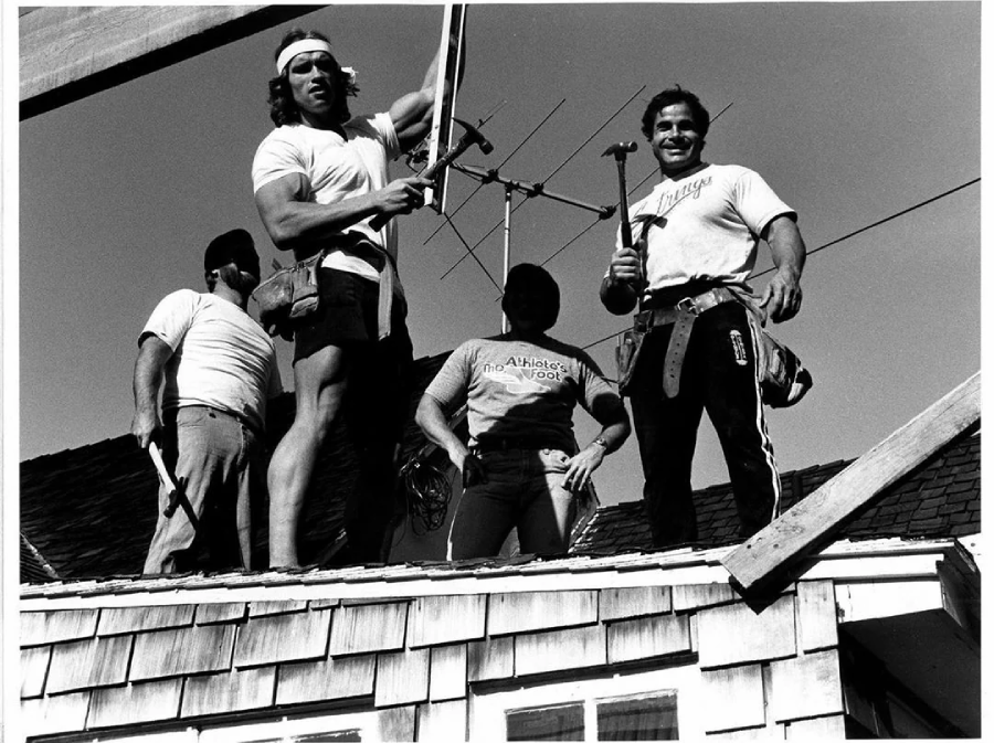 Four men wearing tool belts and holding hammers stand on the roof of a house under construction. Two men face the camera, smiling, while the others work. The scene is bright and outdoors, with a clear sky in the background.