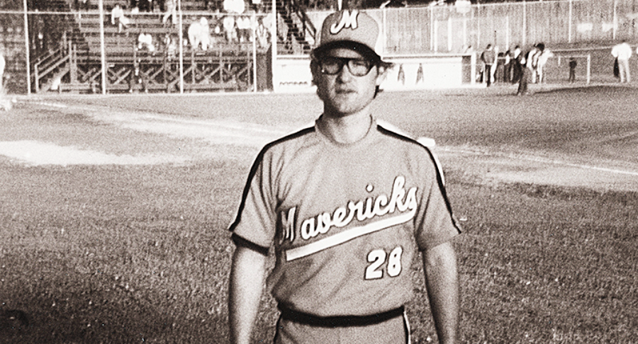 A baseball player in a "Mavericks" uniform with the number 28 stands on a field. The photo is in black and white, and there are people and bleachers visible in the background.