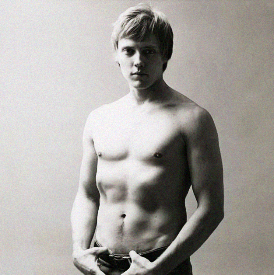 A young man with short light hair stands shirtless against a plain background, looking directly at the camera with a neutral expression. He is holding the waistband of his pants with both hands. The image is in black and white.