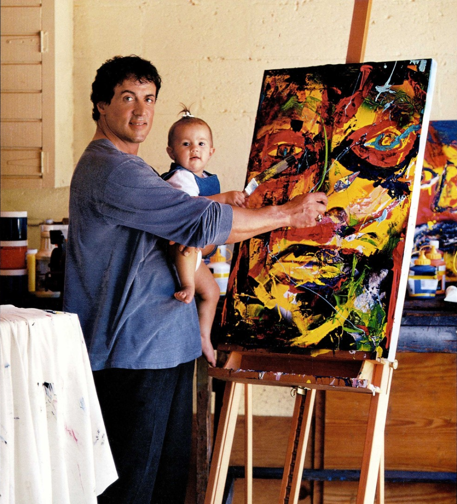 A man holds a baby while painting a colorful, abstract portrait on a canvas set on an easel in an art studio. Art supplies and paint bottles are visible in the background.
