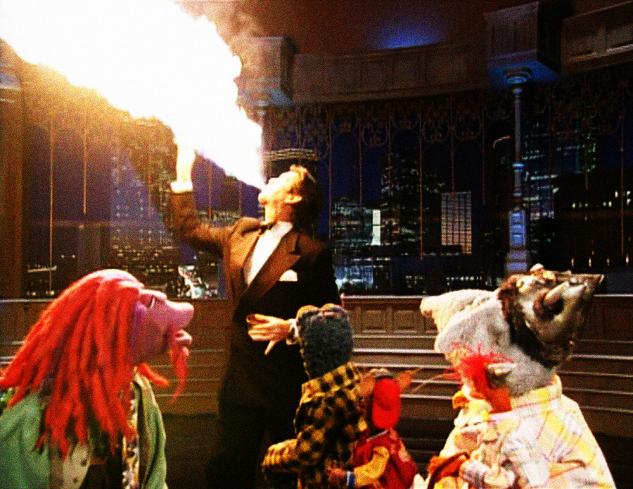 A person in a tuxedo breathes fire on stage while three colorful puppet characters watch in awe against a nighttime cityscape backdrop.