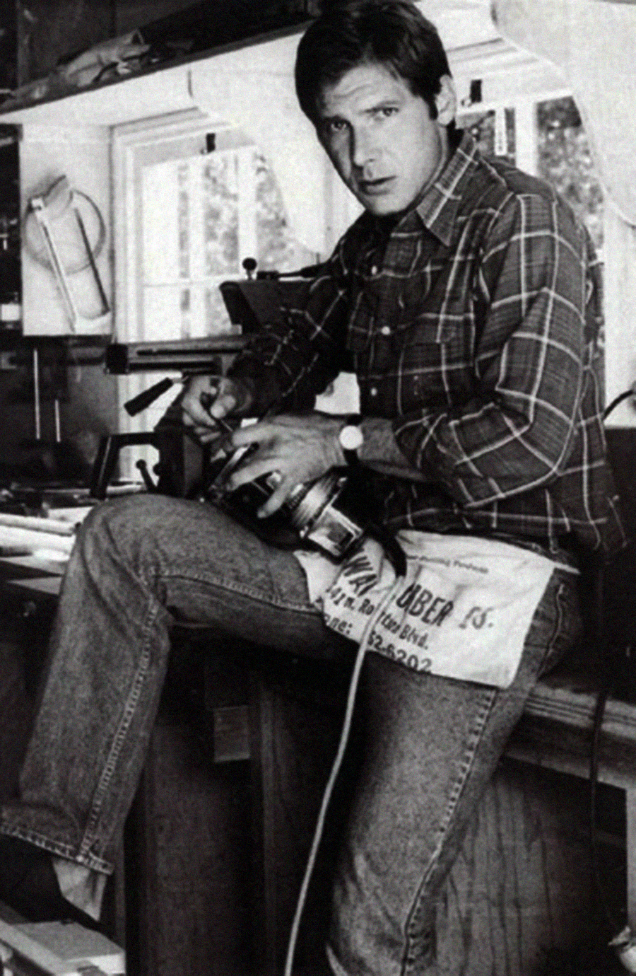 A man in a plaid shirt and jeans sits on a workbench in a workshop, holding a power tool with one hand and wearing a carpenter's apron. He looks toward the camera with a serious expression.