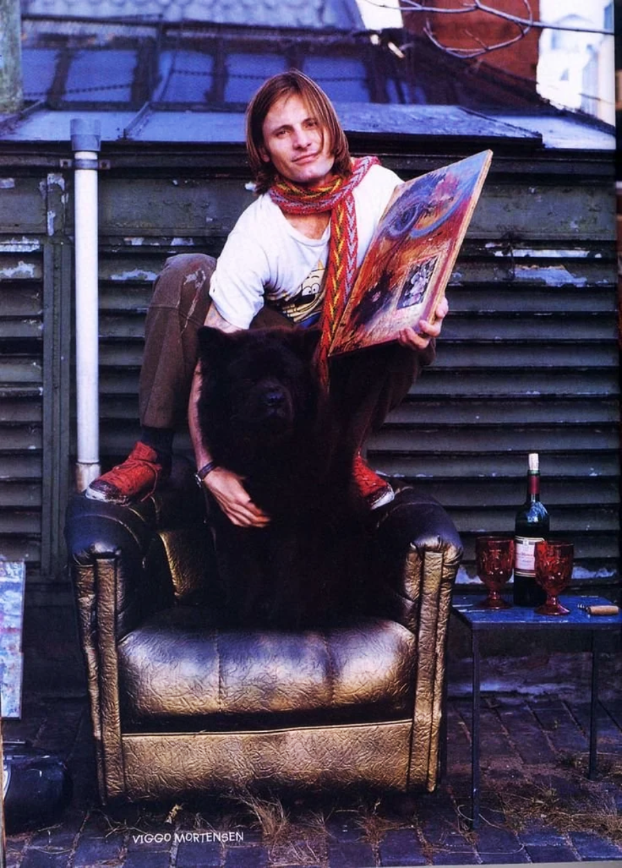 A person with long hair and a scarf sits on the arm of a gold armchair, holding a colorful painting. A large black dog sits in the chair. A wine bottle and two glasses are on the ground nearby, outdoors.
