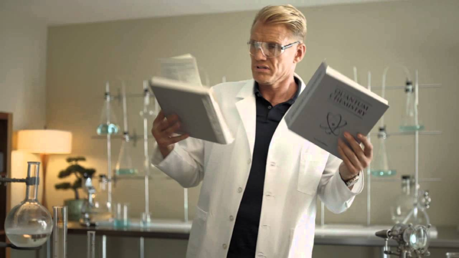 A man in a white lab coat and safety glasses stands in a laboratory, holding and looking confusedly at two large books. Laboratory glassware and equipment are visible on the table and shelves behind him.
