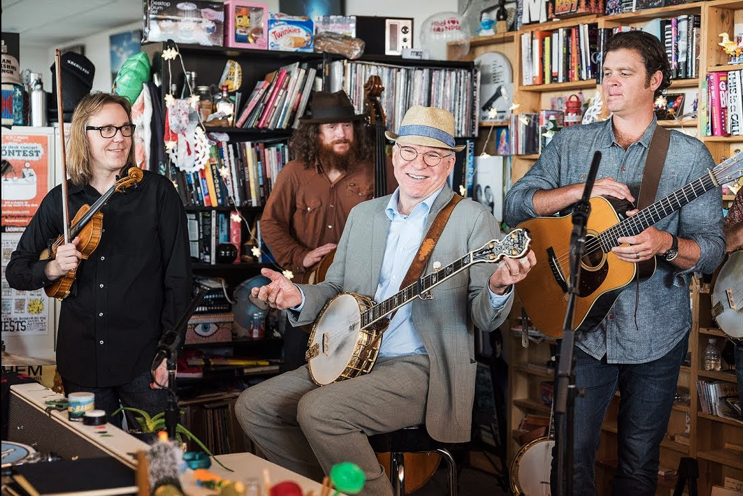Four musicians perform in a cozy, cluttered room full of shelves and books. The seated man in a hat plays banjo and smiles, while the others stand around him holding a guitar, violin, and another string instrument.
