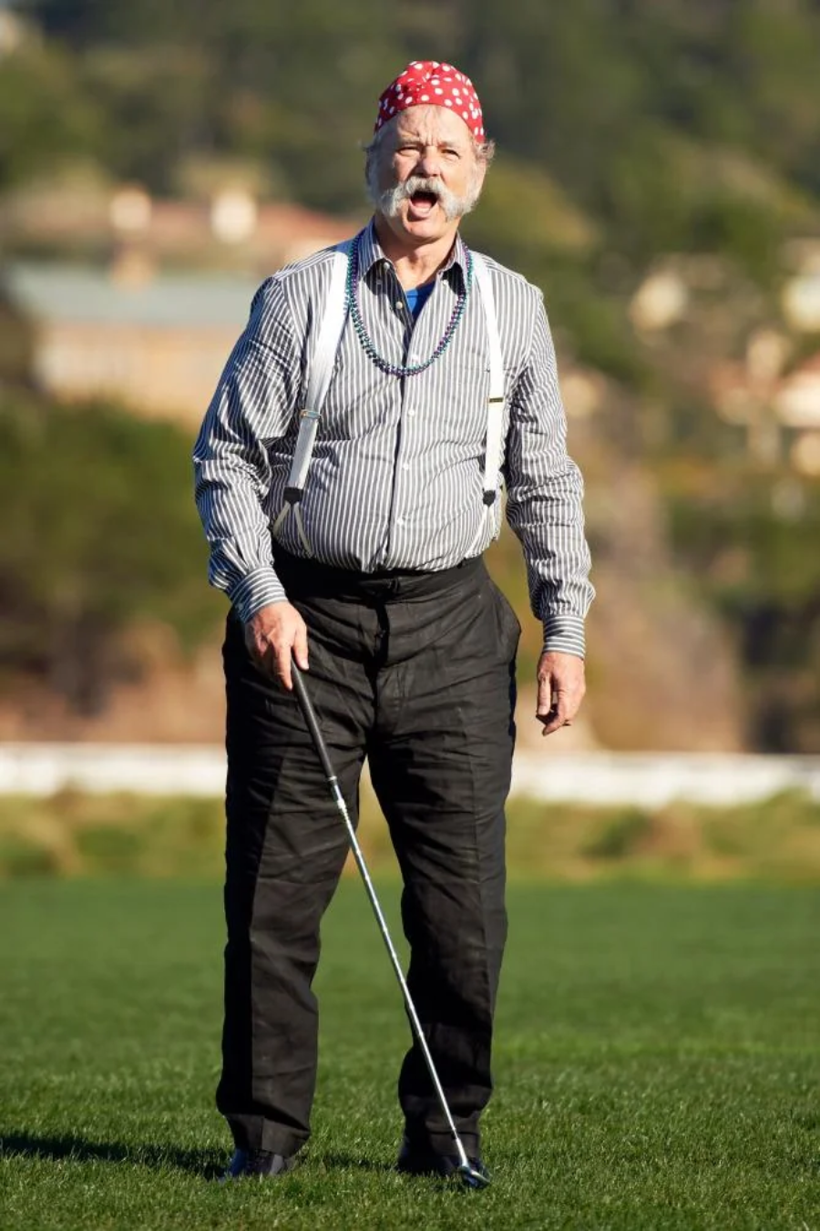 An older man wearing a red polka dot bandana, striped shirt, suspenders, and dark pants stands on a golf course holding a golf club, with an amused expression on his face.