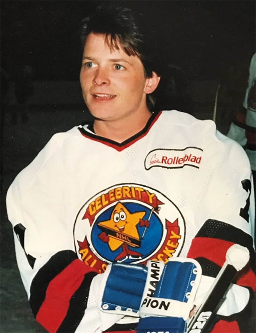 A person wearing a white "Celebrity All-Star Hockey" jersey and blue hockey gloves stands on an ice rink, smiling and looking slightly to the side. The background is dimly lit with other players visible.