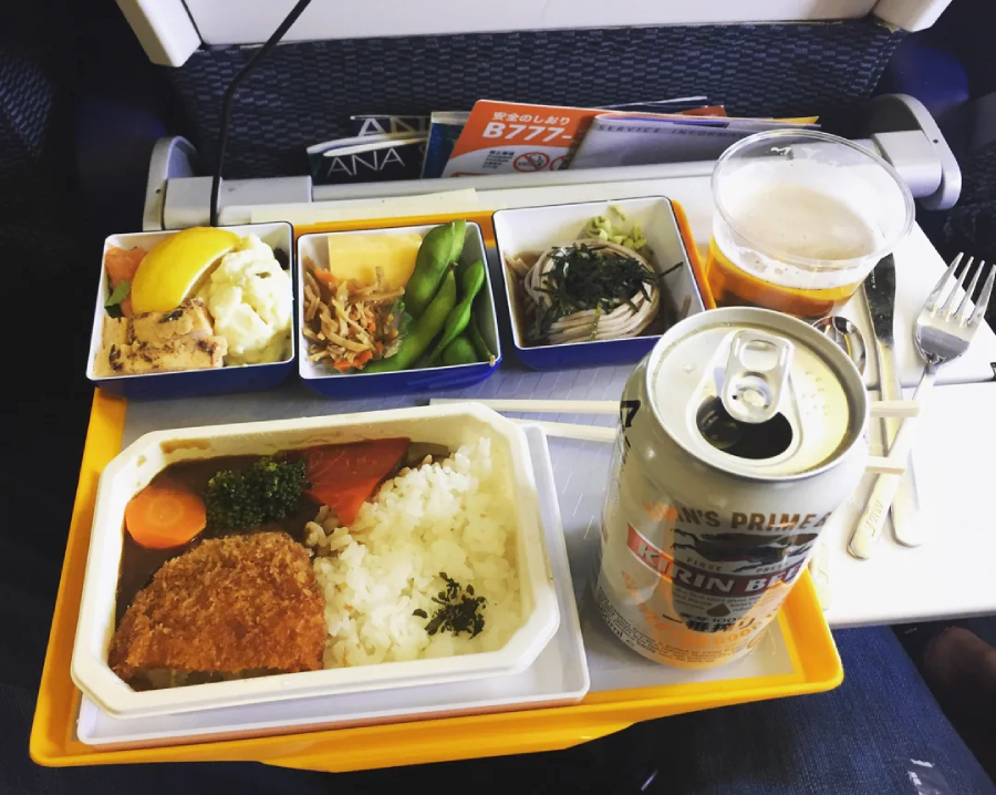 A tray of airplane food with rice, breaded meat, vegetables, sides, a can of Kirin beer, and a glass, placed on a fold-out table in front of an airline seat.