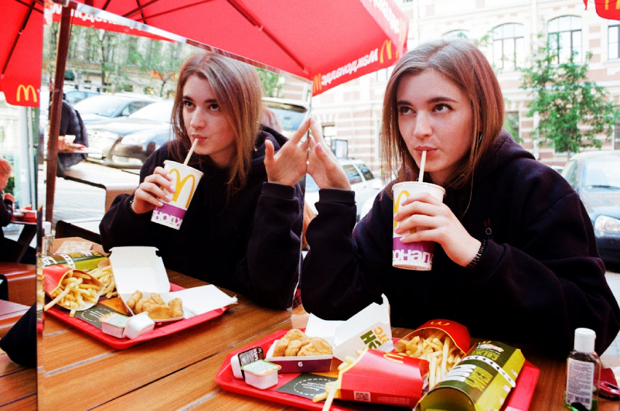 A young woman sits at a McDonald's table with fries, nuggets, and a drink, sipping through a straw while her reflection is visible in a large mirror beside her. Red McDonald's umbrellas are overhead.