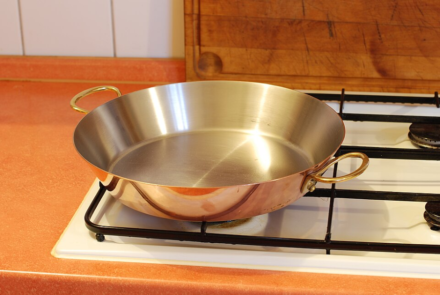 A shiny, empty metal frying pan with two handles sits on a stovetop burner in a kitchen with orange countertops and a wooden cutting board in the background.