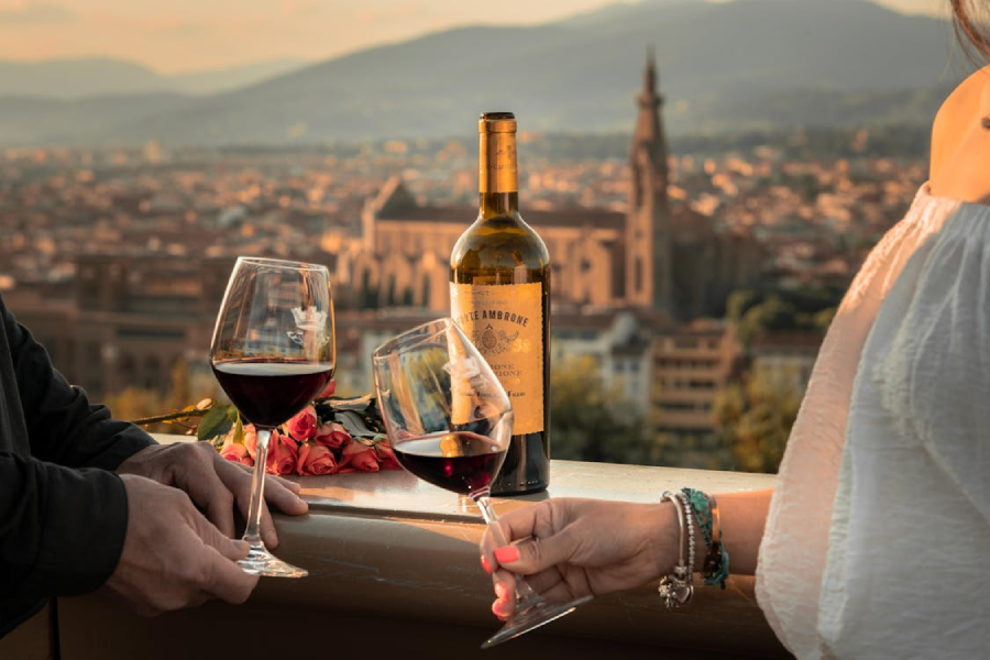 Two people clink glasses of red wine on a terrace overlooking a city at sunset. A wine bottle, bouquet of roses, and a scenic view with a church and mountains are visible in the background.