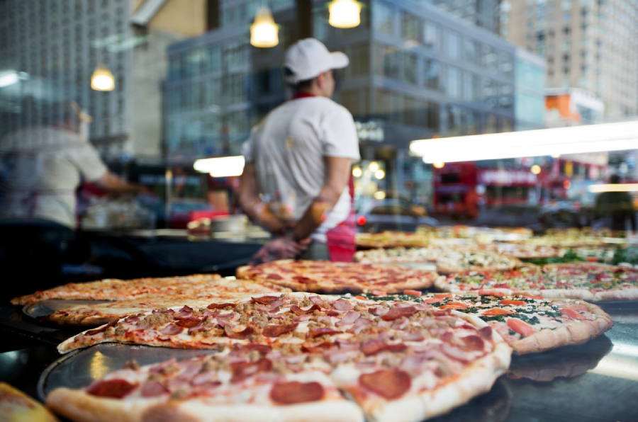 Several large pizzas with various toppings are displayed on a counter in front of a window. A person wearing a white shirt and cap stands behind the counter, city buildings visible outside.