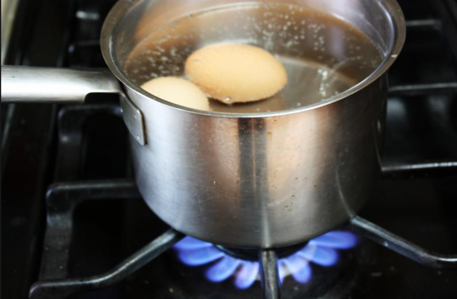 A stainless steel pot with two eggs boiling in water sits on a gas stove with a visible blue flame.
