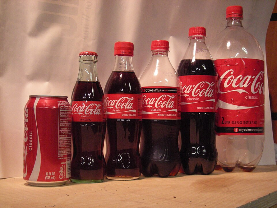 Six Coca-Cola containers of varying sizes and shapes—one can, two glass bottles, and three plastic bottles—are arranged in a row on a flat surface with a neutral background.