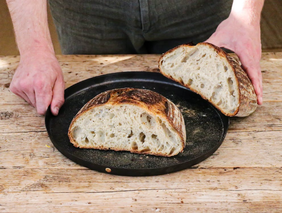 A person holds a round black tray with a rustic loaf of artisanal bread cut in half, showing its airy, textured crumb. The tray rests on a weathered wooden table.