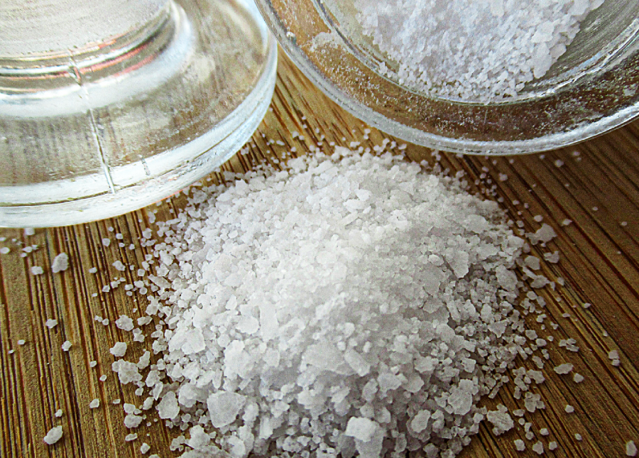 A close-up of coarse sea salt spilled from a glass jar onto a wooden surface, with salt crystals scattered around.