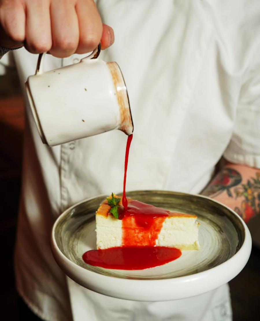A person in a white shirt pours vibrant red sauce from a small white jug onto a slice of cheesecake garnished with a sprig of mint, served on a round ceramic plate.