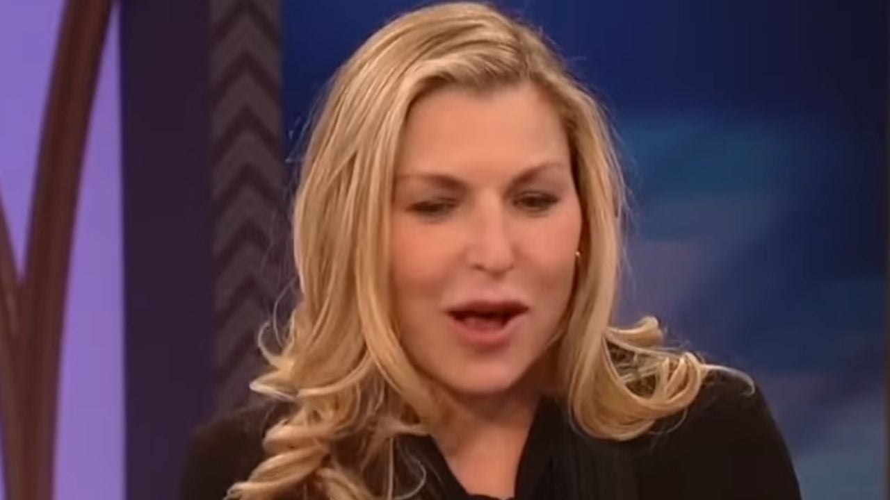 A woman with long blonde hair, wearing a black top, is speaking on a television set with a blurred blue and purple background.