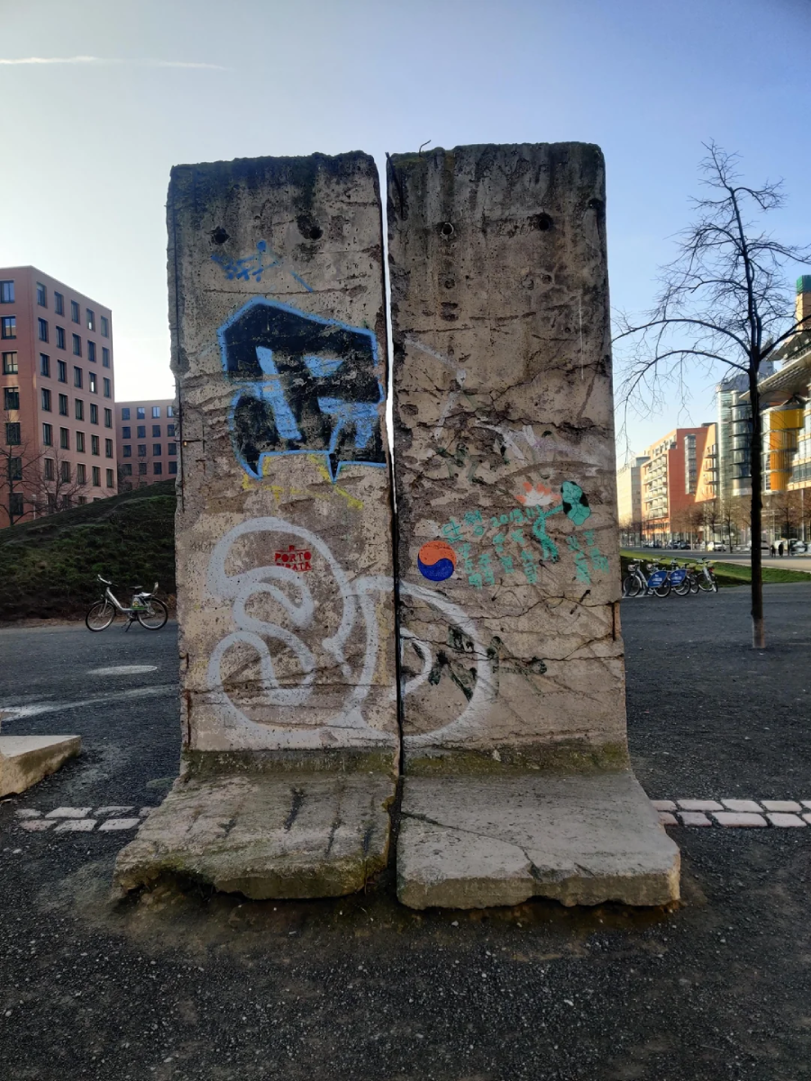 Two large, weathered concrete slabs with graffiti stand outdoors in a city park. Buildings, bicycles, and bare trees are visible in the background under a clear sky.