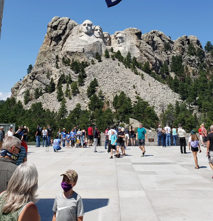 Tourists gather at the viewing area of Mount Rushmore, with the carved faces of four U.S. presidents visible on the mountain in the background under a clear blue sky. Some visitors wear face masks.