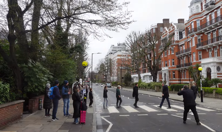 A group of people wait near a crosswalk at a zebra crossing on a city street, while others walk across. Red-brick buildings and trees line the street under an overcast sky.