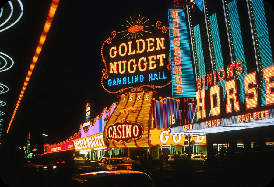 A nighttime street scene in Las Vegas featuring the brightly lit neon signs of the Golden Nugget Gambling Hall and Casino, with classic cars parked out front and vibrant lights illuminating the area.