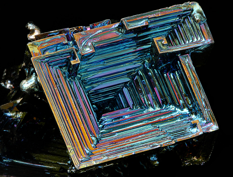 A close-up of a bismuth crystal with an iridescent, metallic surface displaying angular, staircase-like geometric patterns and rainbow colors against a black background.