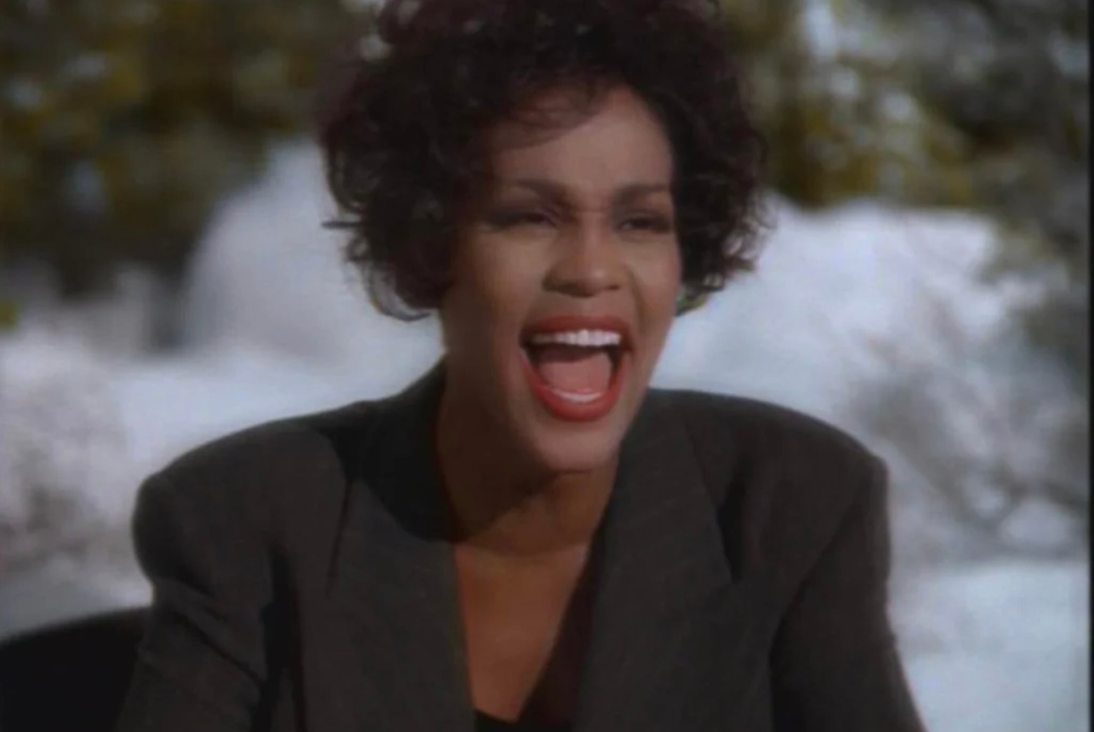 A woman with short curly hair and red lipstick is wearing a dark blazer, smiling broadly and singing outdoors with a snowy background.