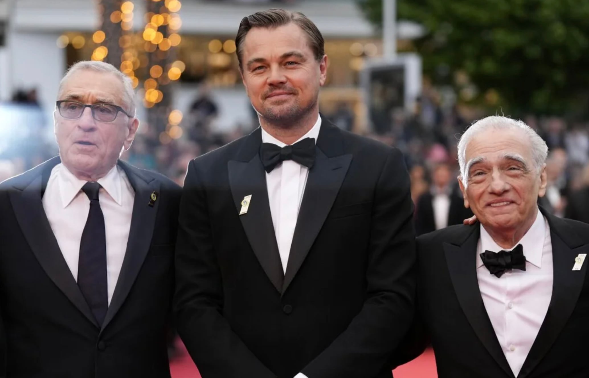 Three men in tuxedos stand together on a red carpet at an event, with blurred lights and trees in the background. They are smiling and appear to be posing for a photo.