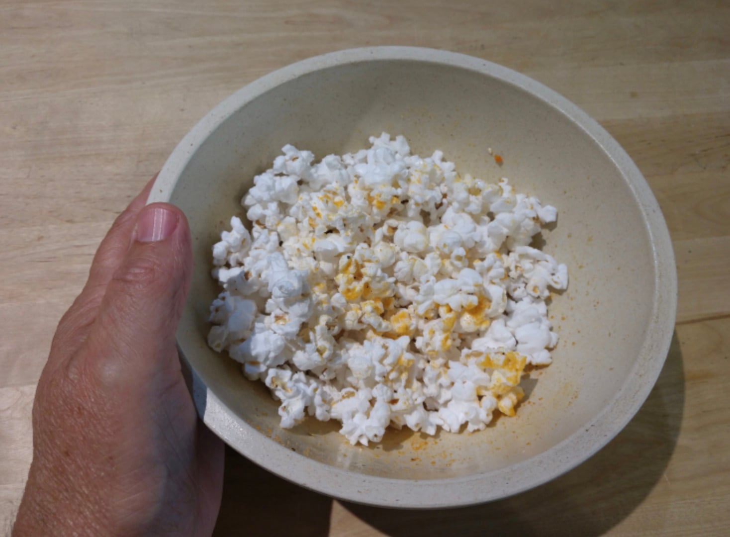 A hand holding a beige bowl filled with popcorn, sprinkled with yellow seasoning, on a light wooden surface.