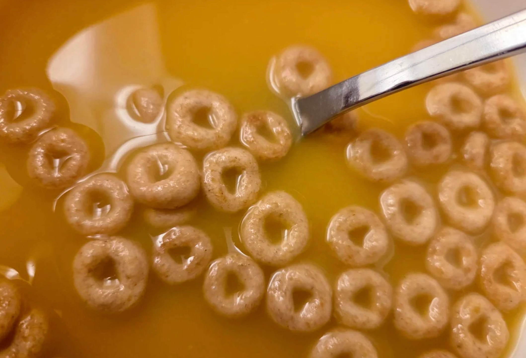 A close-up of round oat cereal floating in orange juice, with a metal spoon partially visible on the right side of the bowl.
