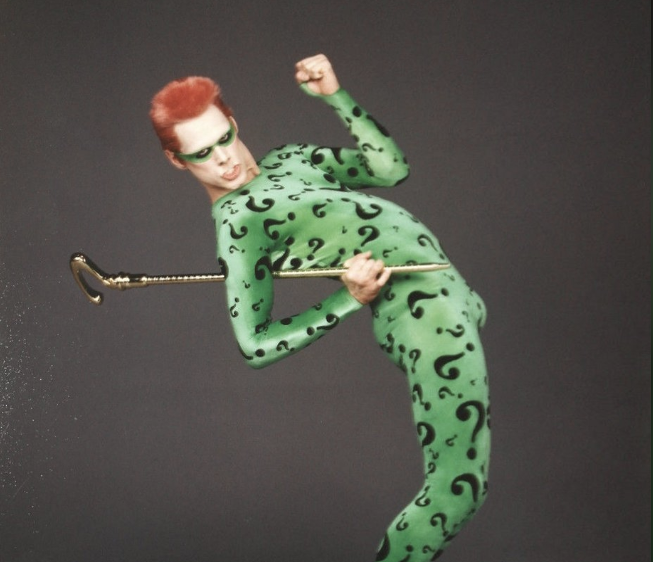 A person in a green bodysuit covered with black question marks strikes a dramatic pose while holding a gold cane. The individual has slicked-back red hair and wears a green mask. The background is plain and dark.